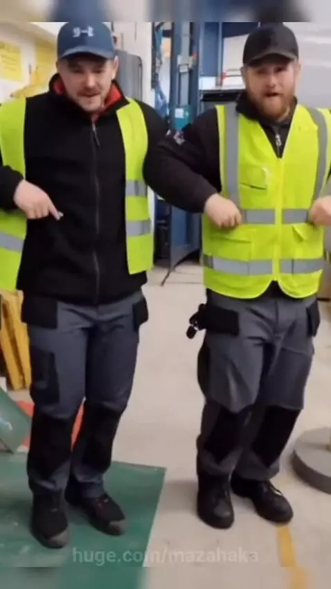 Two men in safety vests and caps enthusiastically dancing in a warehouse setting with industrial equipment.