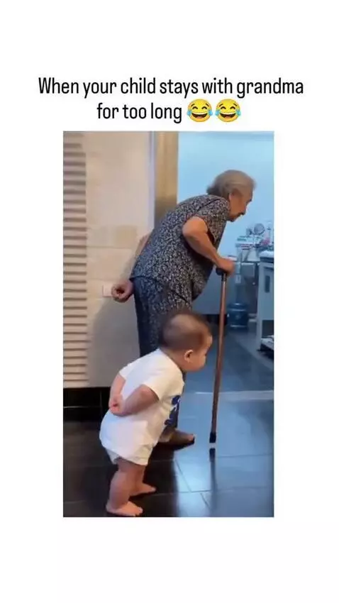 Elderly woman with walking stick and a baby dressed in white, both bent over and walking slowly, mimicking each other.