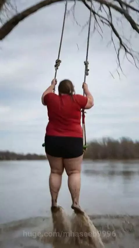 Woman in red shirt and black shorts swinging on a rope swing over a muddy river before splashing in.