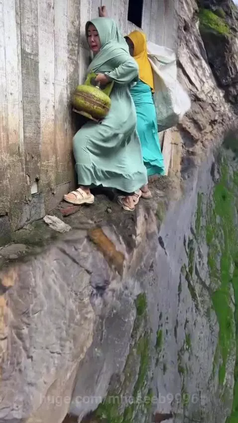 Two women in hijabs and dresses on a narrow ledge with a green valley below, holding a gas cylinder.