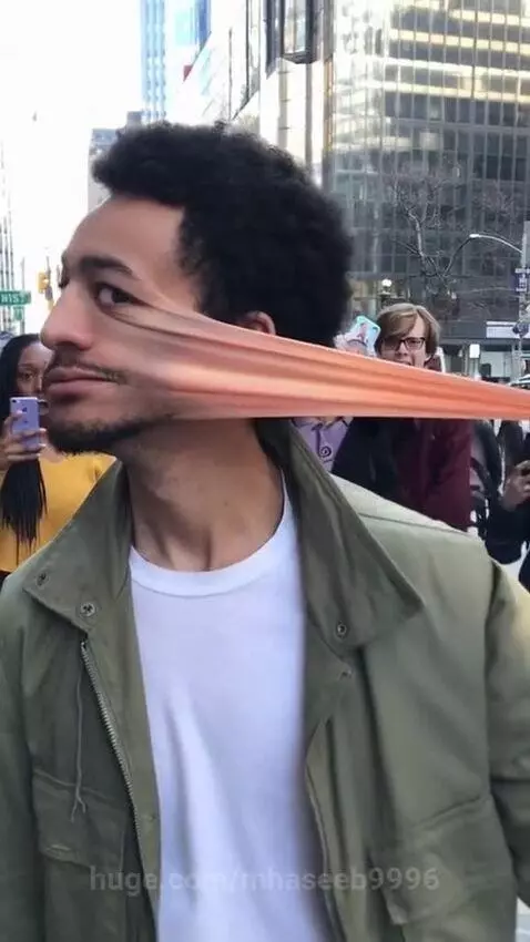 Man with curly hair and beard stretching his cheek skin out from his face on a city street.