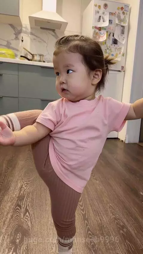 Toddler in pink shirt and leggings performing high kicks and balancing in a home kitchen.