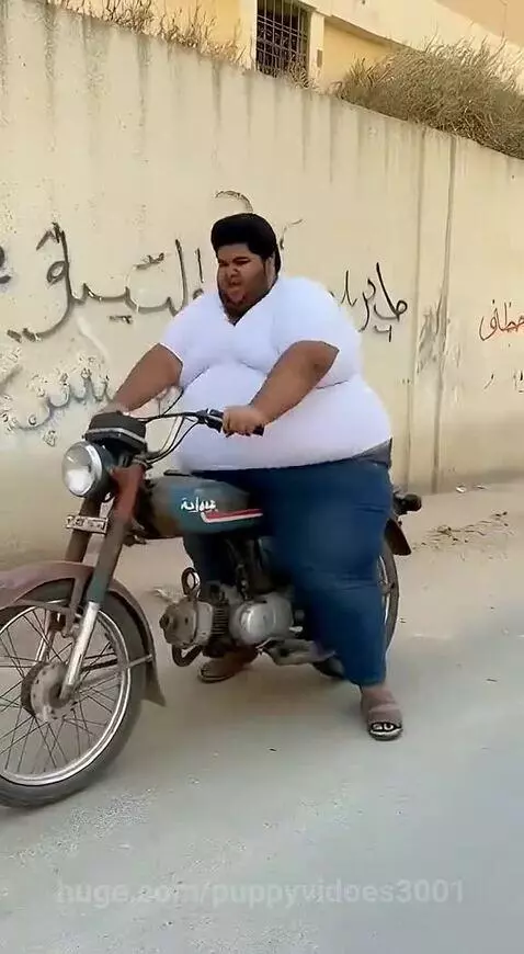 Large man in white shirt and blue jeans falls off a small motorcycle next to graffiti wall.
