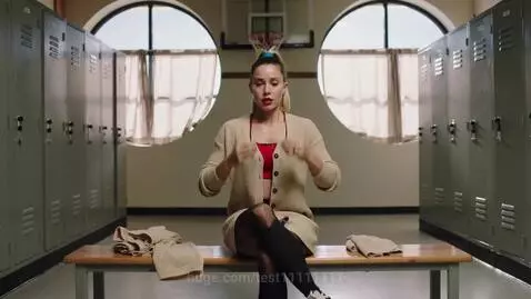 Woman in a locker room changing into a cheerleader outfit, wearing a red sports bra and pleated skirt.