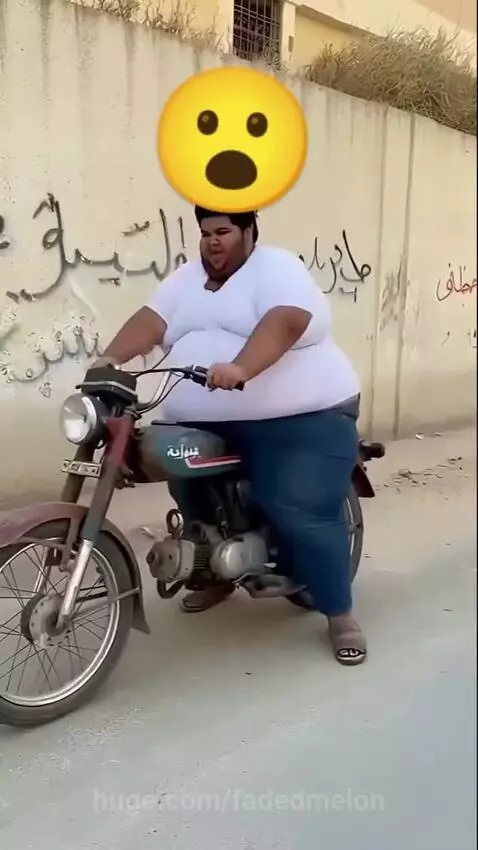 Overweight man in white t-shirt and blue jeans falls off a small motorcycle after attempting to rev the engine.