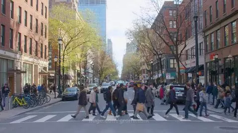 Wide shot of a sunny city street with many pedestrians crossing and walking on sidewalks, cars driving, and buildings with shops.