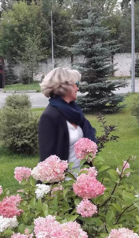 Woman with blonde hair and glasses smiling, standing near pink and white hydrangeas, then walking towards a festive outdoor party setup.