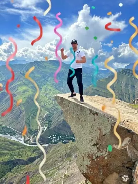 Man dancing on a mountain cliff edge with confetti, overlooking a valley. Russian 'Happy Birthday Anton' banner visible.