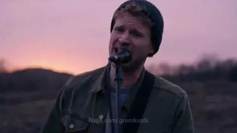 Man with beanie singing passionately into microphone outdoors with a beautiful sunset and hills in the background.