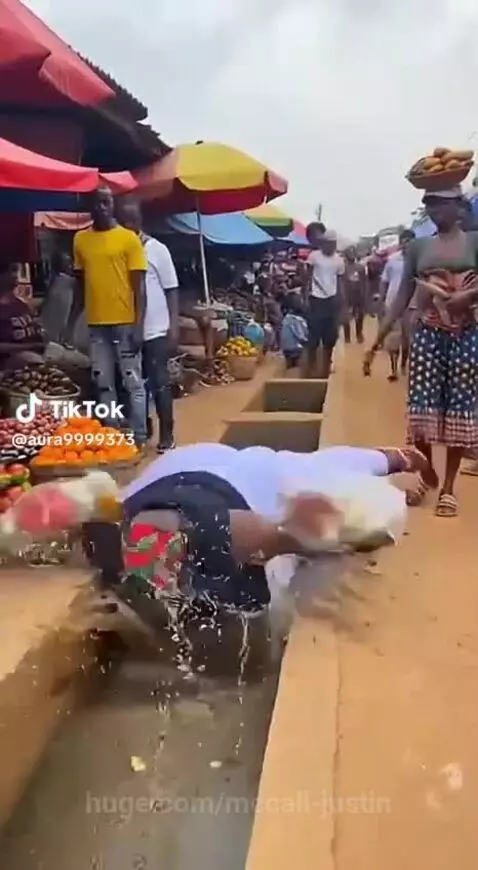 Woman falling into a dirty water ditch after a wooden plank breaks in a busy outdoor market.
