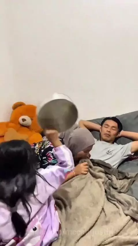 A girl places a metal bowl over a man's blanket-covered foot while a woman watches, anticipating a prank.