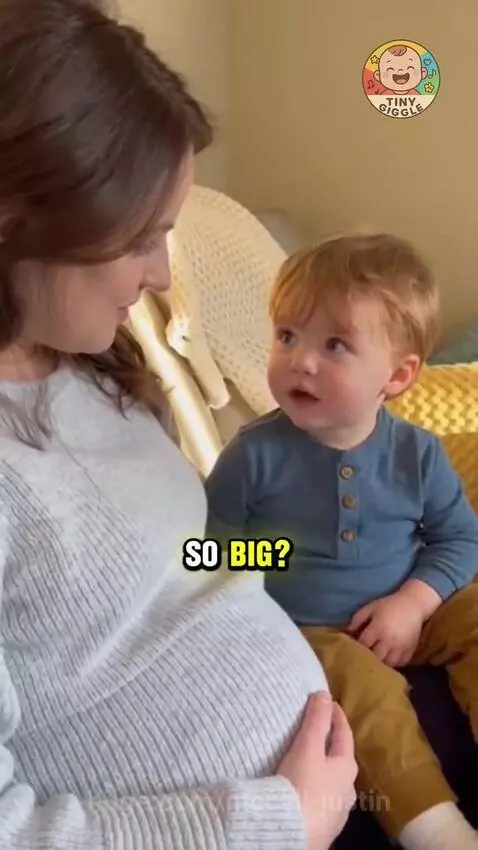 Young boy with red hair looking up at his pregnant mother's belly with wide eyes, asking a funny question.