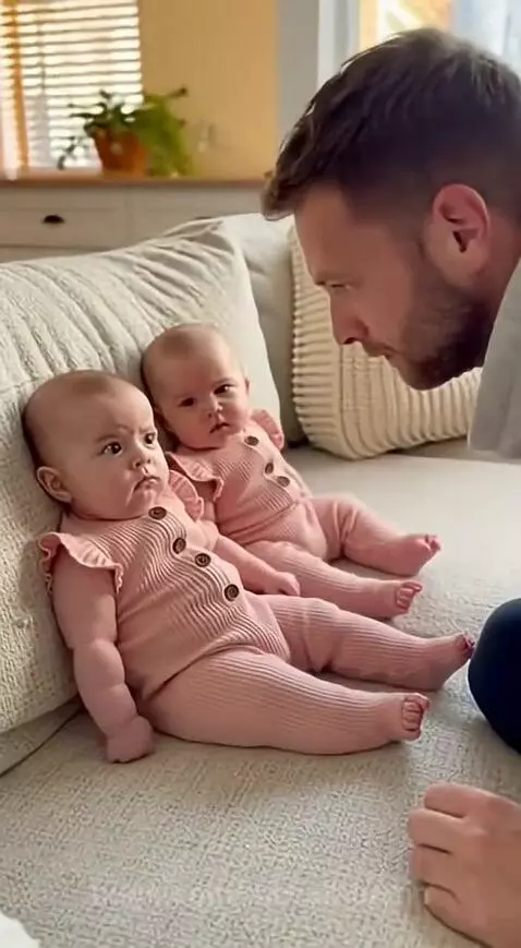 Two infant twin babies in pink rompers on a couch, one mimicking a sneeze sound while their dad laughs.