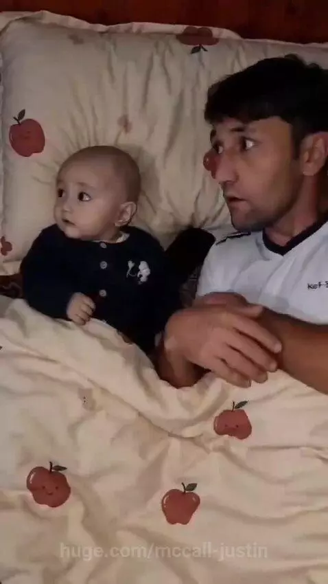 A baby lying in bed with a man, smiling and laughing as the man pretends to sleep under a blanket.