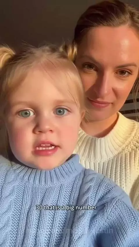 Young girl with blonde pigtails looks at camera, her mom laughs behind her.