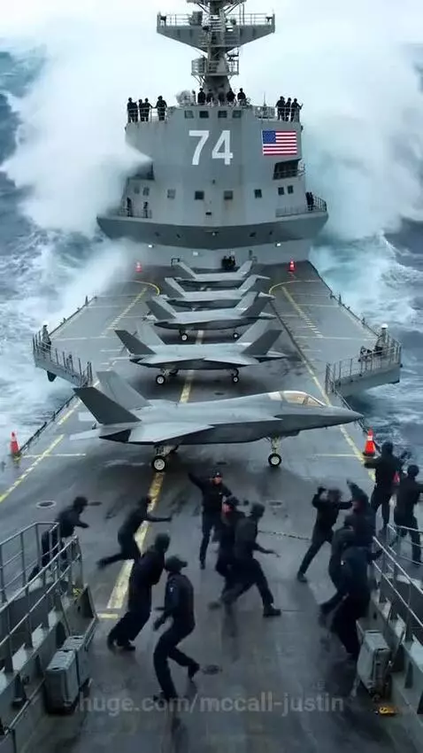 US naval ship with fighter jets on deck battling massive waves during a storm.