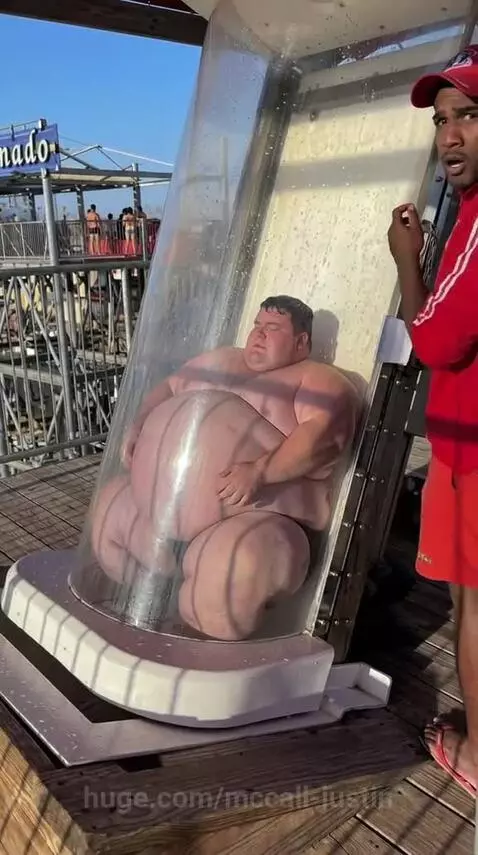 Large man stuck in a clear water slide tube, lifeguard assisting.
