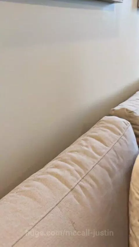 Orange tabby kitten peeking from behind a couch to surprise a man sitting on it.