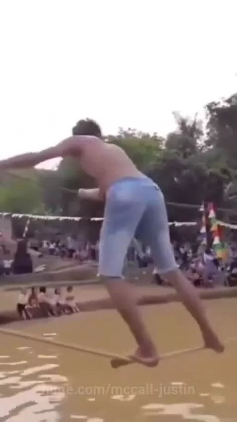 Shirtless man in shorts falls face-first into muddy water while trying to balance on a rope with a stick.