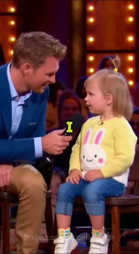 Young girl with blonde hair in a yellow sweater being interviewed by a host on stage.