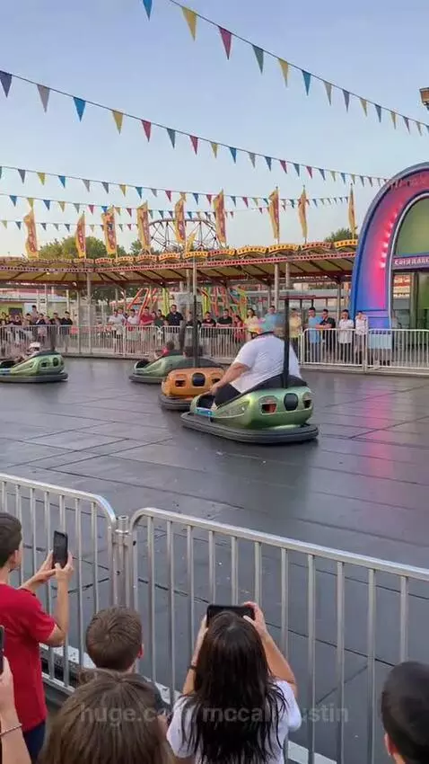 A large man wearing a white t-shirt, black shorts, and a colorful helmet drives a green bumper car at an amusement park, smiling.