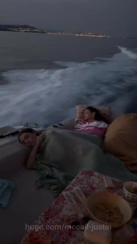 Three women sleeping on cushions on a boat at dusk, with mountains and coastal town lights in the background.