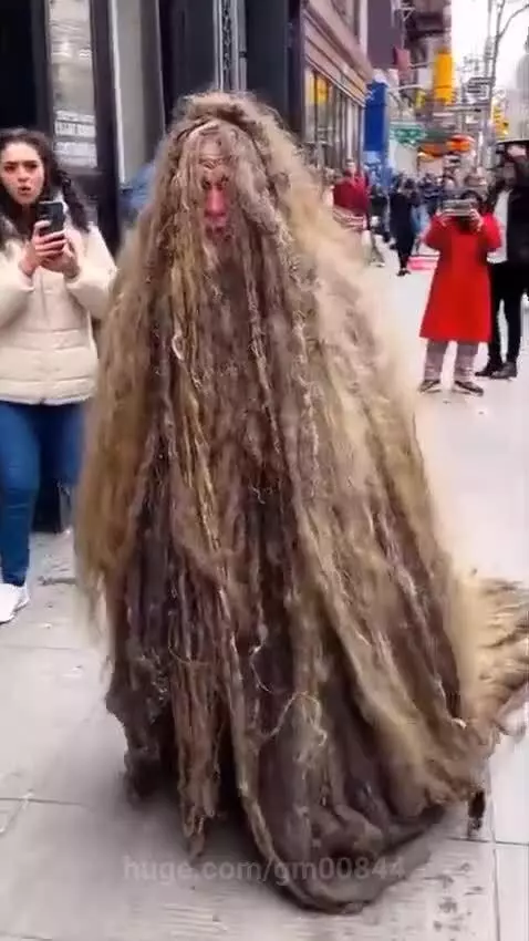 Person in a full-body shaggy hair costume walking down a city sidewalk, with onlookers watching in surprise.