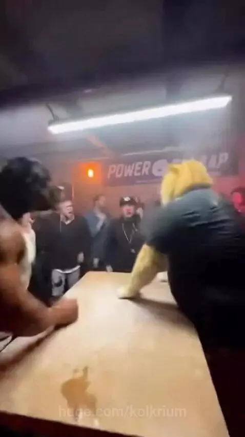 Cat-headed character roaring aggressively at the camera after losing an arm wrestling match to a dog-headed character.