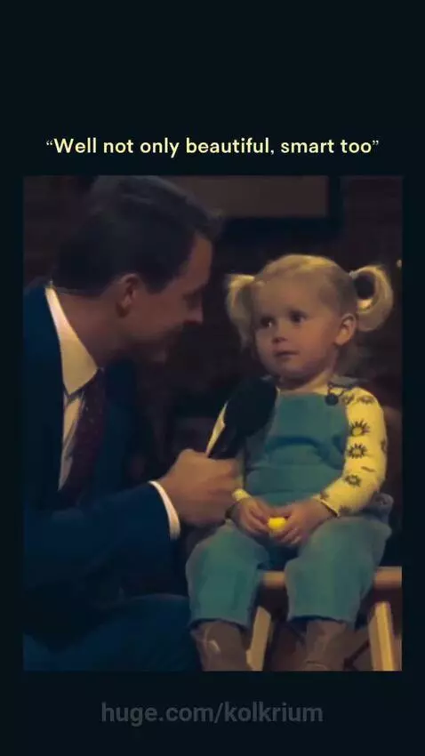 A young girl with pigtails in overalls smiles sweetly while being interviewed by a man in a suit.