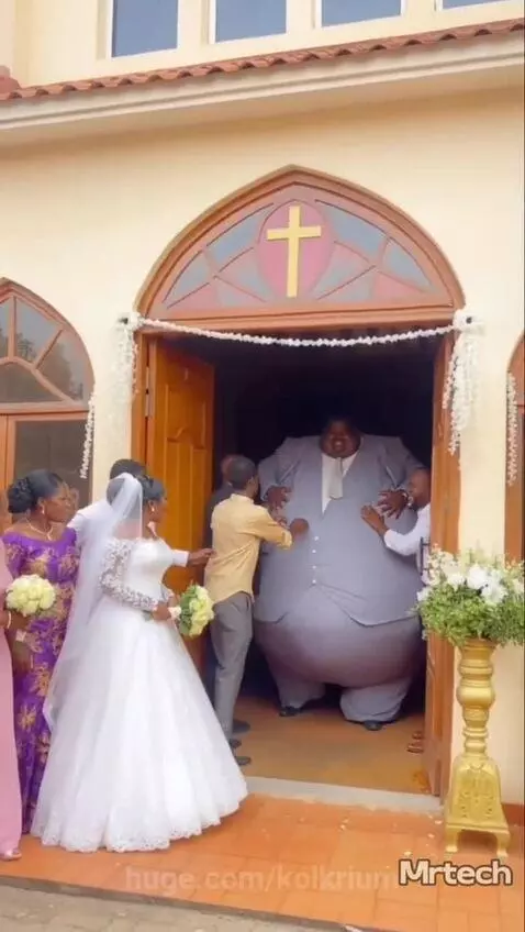 Comedic scene of a very large groom in a padded suit being helped through a church doorway by wedding guests.