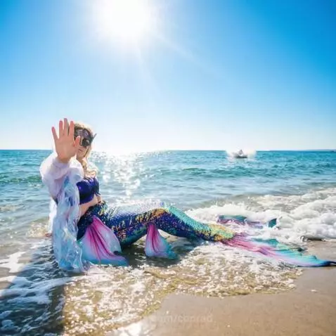Mermaid with iridescent tail sitting on a sandy beach, a whale breaches in the ocean behind her.