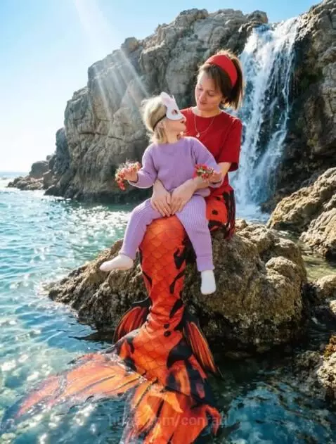 Woman in red top and mermaid tail with child in bunny mask on lap, by a waterfall and blue sea.