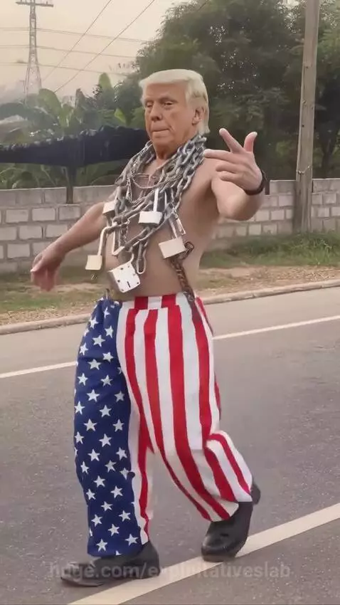 Man with blonde wig resembling Trump, shirtless, wearing American flag pants and multiple chains, walking.