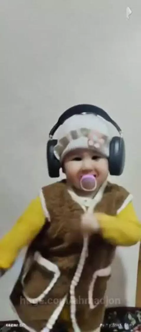 Baby wearing headphones and a pacifier, happily dancing and clapping in front of a light-colored wall.