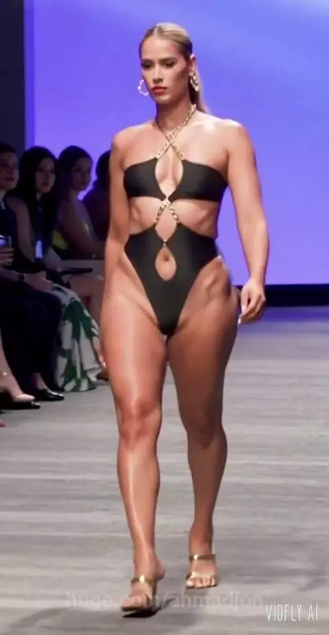 Blonde model in a black strapless swimsuit with gold chain details walks a runway.