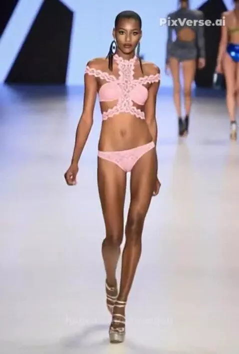 Fashion model with dark skin and short hair walks down a runway wearing a pink lace lingerie set and silver platform heels.
