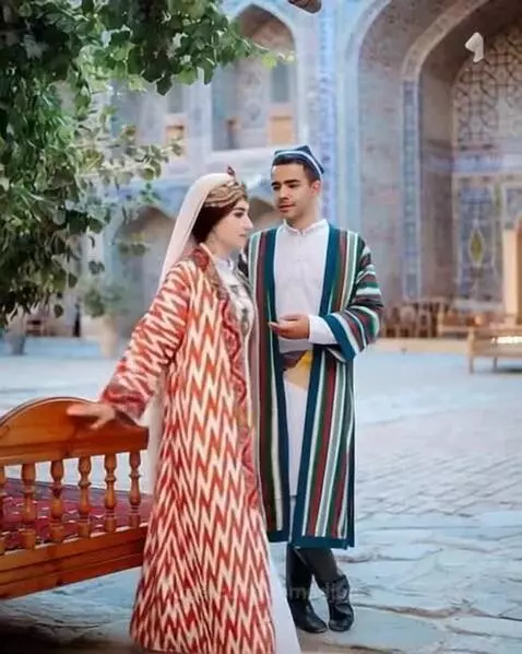 Uzbek bride and groom in colorful traditional wedding attire, standing in an ornate tiled courtyard.