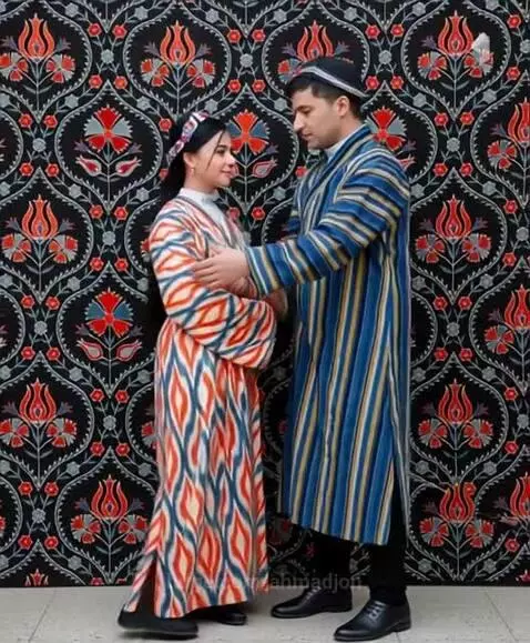 Couple in traditional Central Asian attire, woman in ikat dress, man in striped coat, against floral patterned wall.