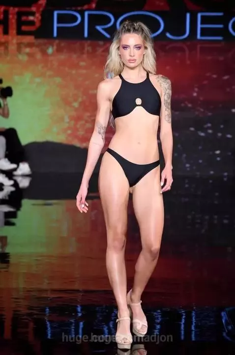 Female model with blonde hair and blue streaks walks a runway in a black bikini, posing confidently.