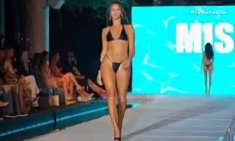 Model with long hair walks a runway in a black bikini and high heels, audience in background.