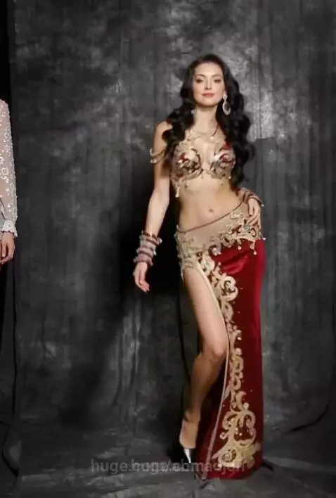 Woman in elaborate red and gold belly dance costume posing against a dark textured background.