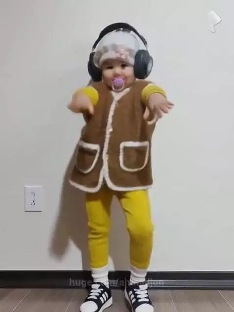 A toddler wearing a white hat, large headphones, and a fuzzy vest dances energetically in front of a white wall.