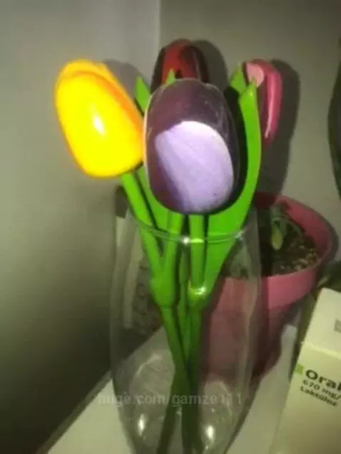 Close-up of a clear glass vase holding four vibrant artificial tulips: yellow, purple, red, and pink, with green stems and leaves.