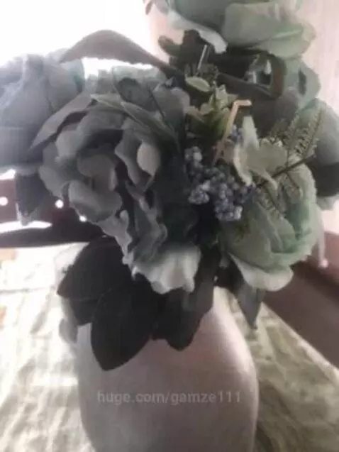 A bouquet of artificial light green and blue flowers with dark green leaves in a light-colored vase.