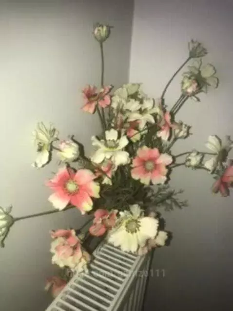 A bouquet of pink and white artificial flowers with green leaves arranged on a white radiator in a room.