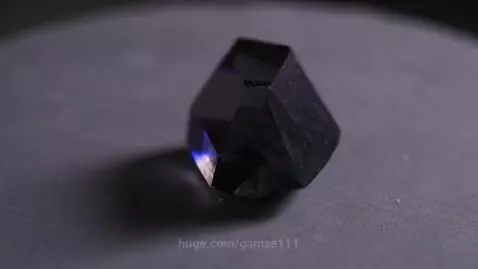 Close-up of a dark, faceted crystal with a subtle blue light reflection on a grey surface.