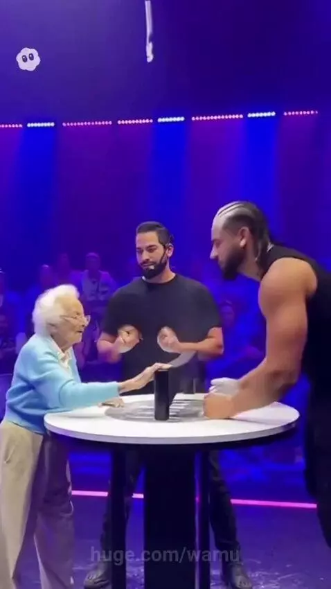 Elderly woman in blue cardigan falls backward after a slap challenge, glasses off, on stage.