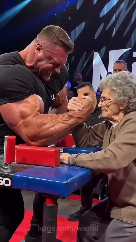 A muscular man with prominent veins arm wrestling an elderly woman with glasses, both smiling.