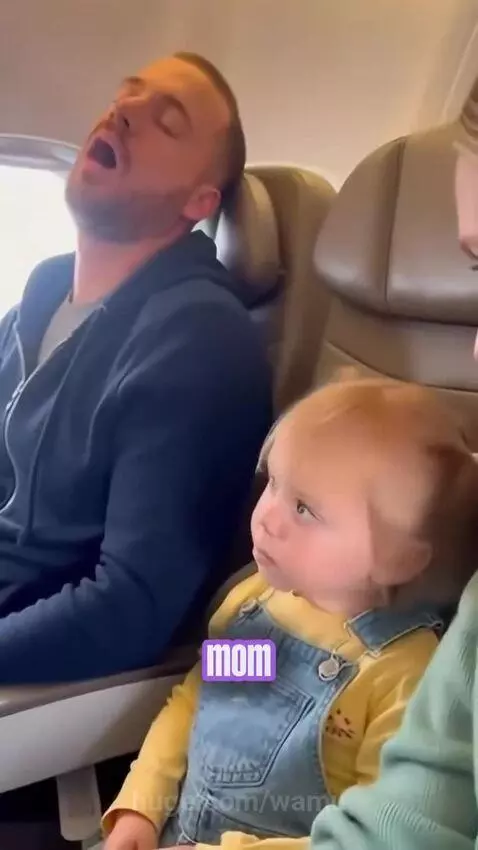 A young girl with pigtails looks seriously at the camera next to a man snoring loudly on an airplane.