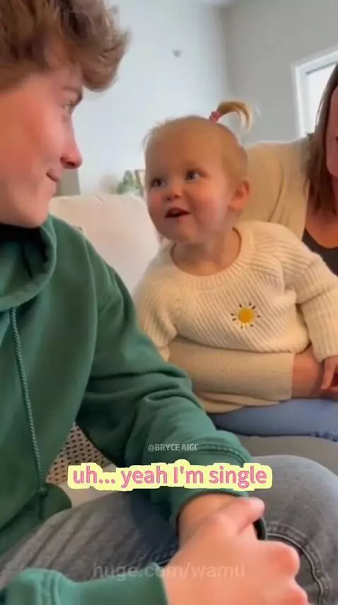 Man in green hoodie smiling at a baby girl with pigtails wearing a sun sweater, who is sitting on an adult's lap.